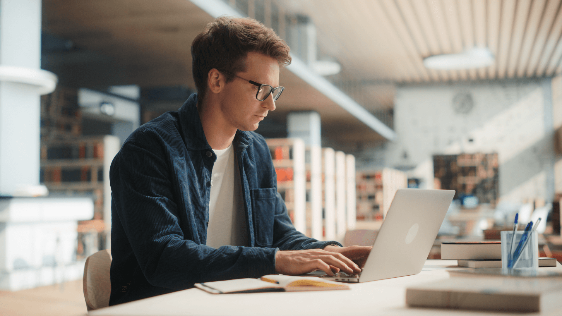 A young man wearing glasses works on a laptop at a desk in a modern library, with bookshelves and soft lighting in the background. An open notebook and a glass with pens are on the table beside him.